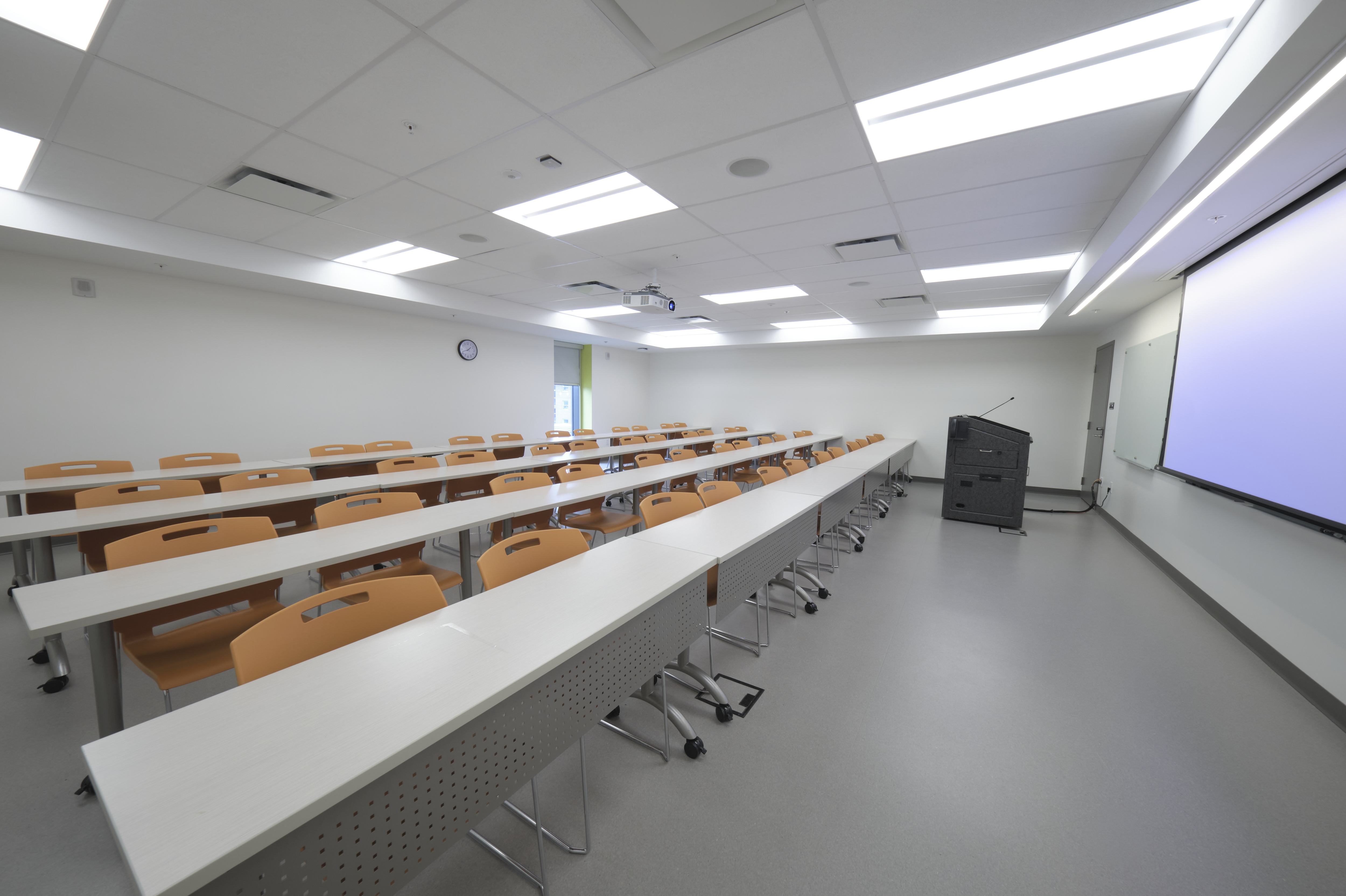 A room with rows of tables and chairs with a lectern, whiteboard and projector screen at the front.