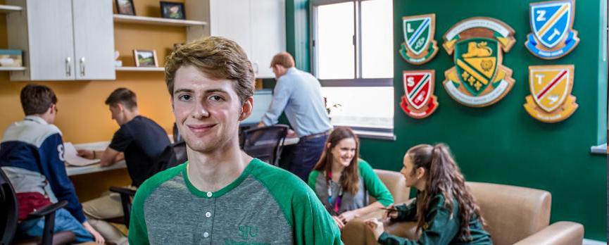 Matteo, a white male presenting person, stands in foreground of the colourful Experience Centre. Other ambassadors stand behind him and on the wall are the House crests surrounding the SJU official crest.