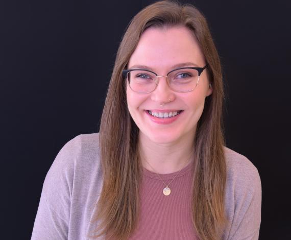 Angelina, a white femme presenting person with long brown hair, green eyes, smiles in front of a beige background