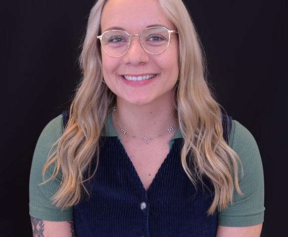 Britt, a white femme presenting person with long blonde hair, smiles in front of a black background