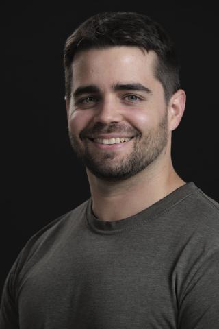 Carl, a masculine presenting person with dark hair and a short beard, smiles in front of a black background.